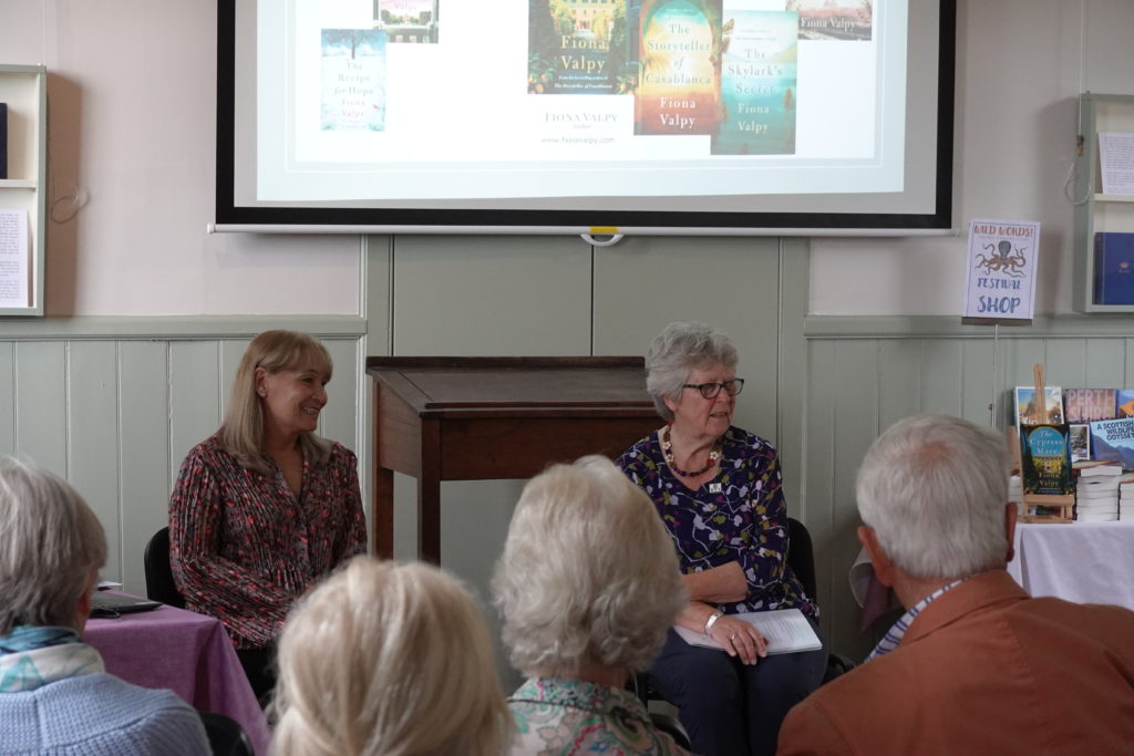 Author Fiona Valpy sitting with library volunteer Gillean Ford, in front of powerpoint presentation showing covers of Valpy's books.