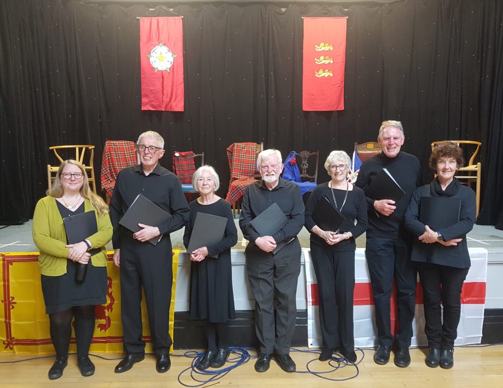 Image shows seven people wearing black, standing in front of a stage, holding script books.