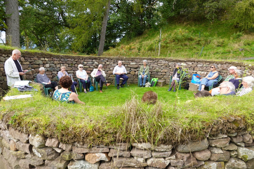 Sixteen people sitting inside a stone storytelling circle, with grass atop and in the middle.